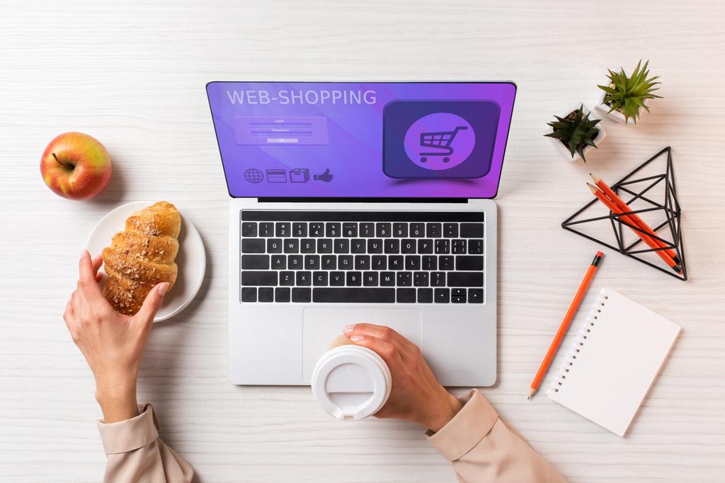 abgeschnittene Aufnahme einer Geschäftsfrau, die Einweg-Kaffeetasse und Croissant hält, während sie Laptop mit Web-Shopping-Anwendung benutzt - Foto, Bild