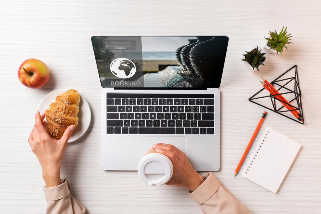 cropped shot of businesswoman holding disposable coffee cup and croissant while using laptop with booking website  - Photo, Image