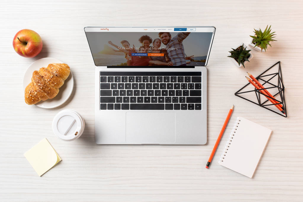 vista superior do laptop com site de couchsurfing, croissant, maçã e xícara de café descartável na mesa de escritório
    - Foto, Imagem