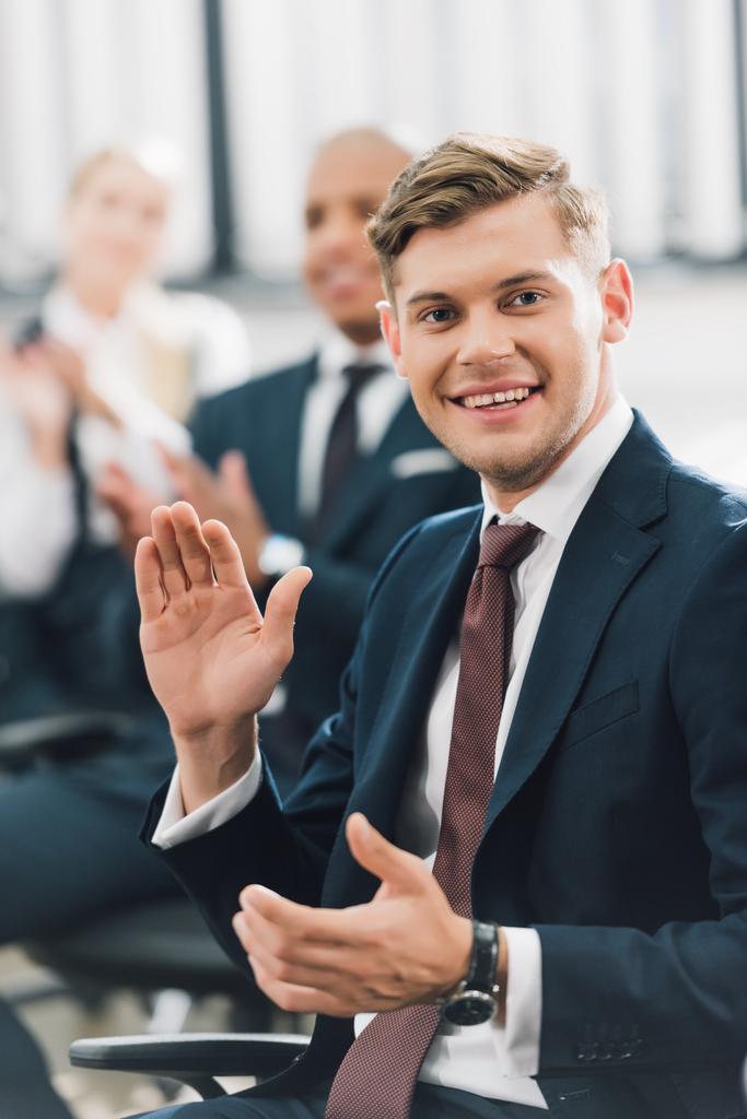 onnellinen nuori liikemies heiluttaa kättä ja hymyilee kameralle toimistossa
 - Valokuva, kuva