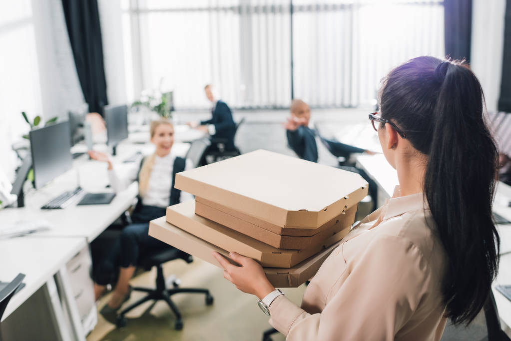 mujer joven sosteniendo cajas de pizza y mirando a sus compañeros de trabajo en la oficina
 - Foto, imagen