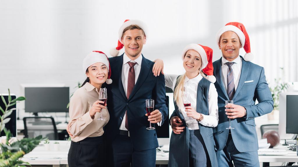 onnellisia nuoria kollegoita jouluhatuissa juomassa viiniä ja hymyilemässä kameralle juhliessaan uutta vuotta toimistossa
  - Valokuva, kuva