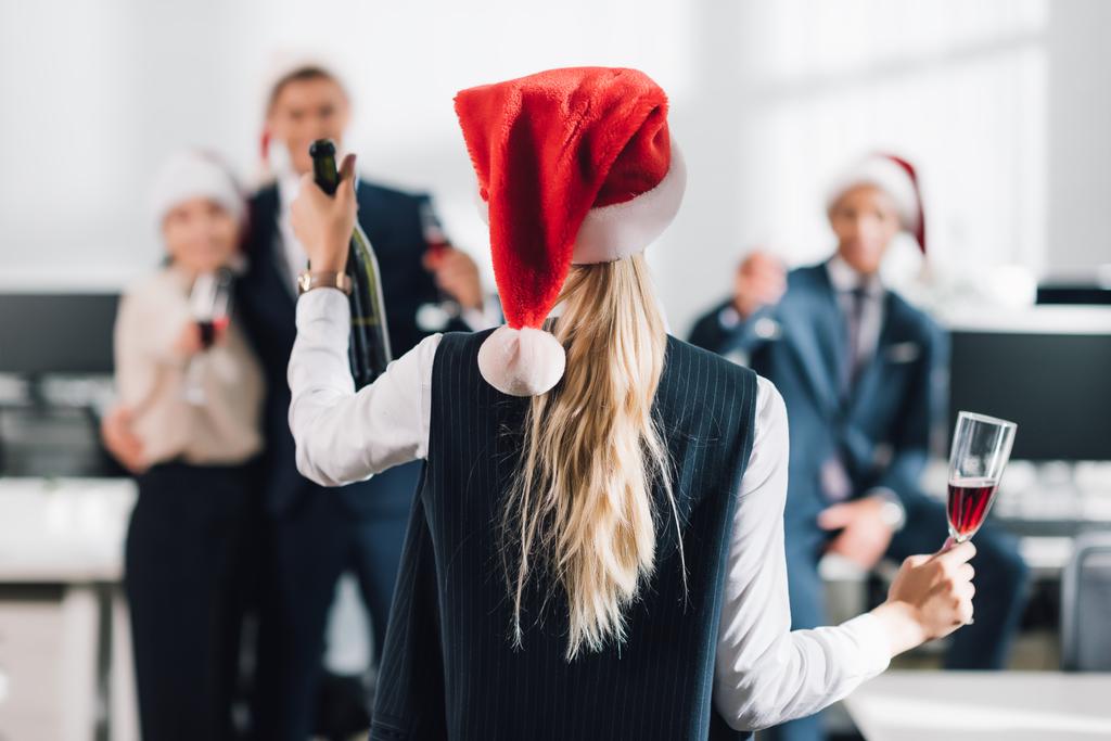 Rückansicht einer Geschäftsfrau mit Weihnachtsmann-Hut, die Glas und Flasche Champagner hält, während sie mit Kollegen Weihnachten feiert - Foto, Bild