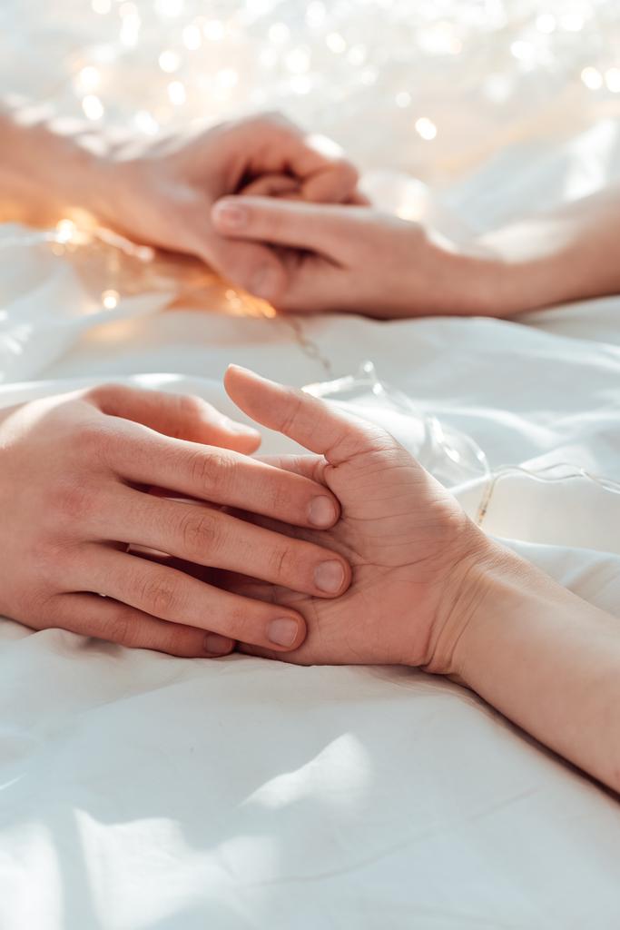 ガーランドと一緒にベッドで寝ながら手を繋いでいるカップルの部分的なビュー - 写真・画像