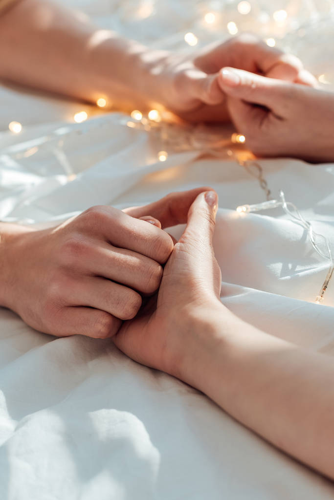 visão parcial do casal de mãos dadas enquanto deitado na cama com guirlanda
 - Foto, Imagem