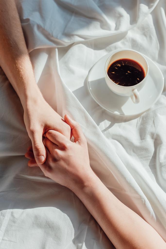 colpo ritagliato di coppia che si tiene per mano su lenzuolo bianco con tazza di caffè aromatico
 - Foto, immagini
