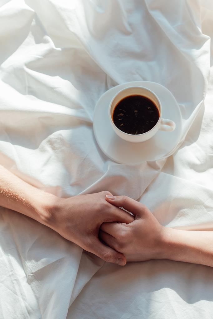 обрізаний знімок пари, що тримає руки на білому простирадлі з чашкою ароматної кави
 - Фото, зображення