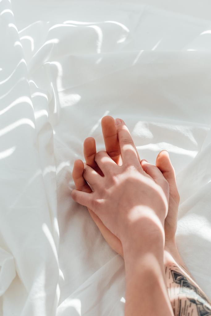 vista parziale di coppia amorevole che si tiene per mano mentre sdraiato su lenzuolo bianco
 - Foto, immagini