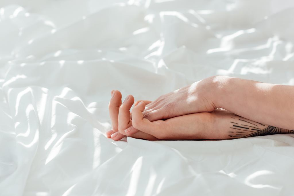 partial view of loving couple holding hands while lying on white bed sheet - Photo, Image