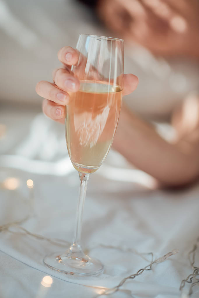 partial view of woman holding glass of champagne on white bed sheet with garland - Photo, Image