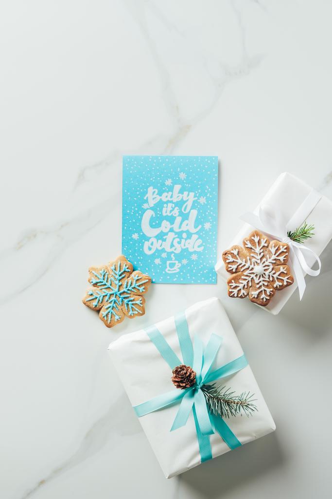 Draufsicht auf weiße Weihnachtsgeschenke, Schneeflockenkekse und Grußkarte mit "Baby seine Kälte draußen" Schriftzug auf Marmortisch - Foto, Bild