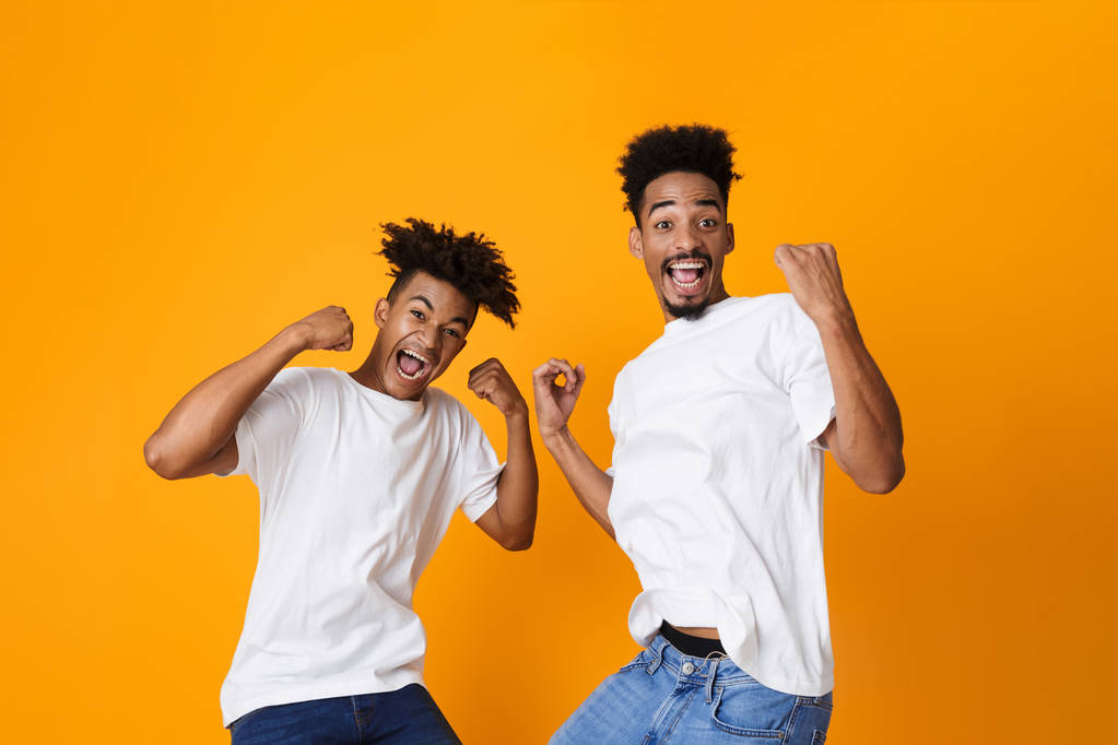 Two excited male african friends in t-shirts standing isolated over yellow background, celebrating success - Photo, Image