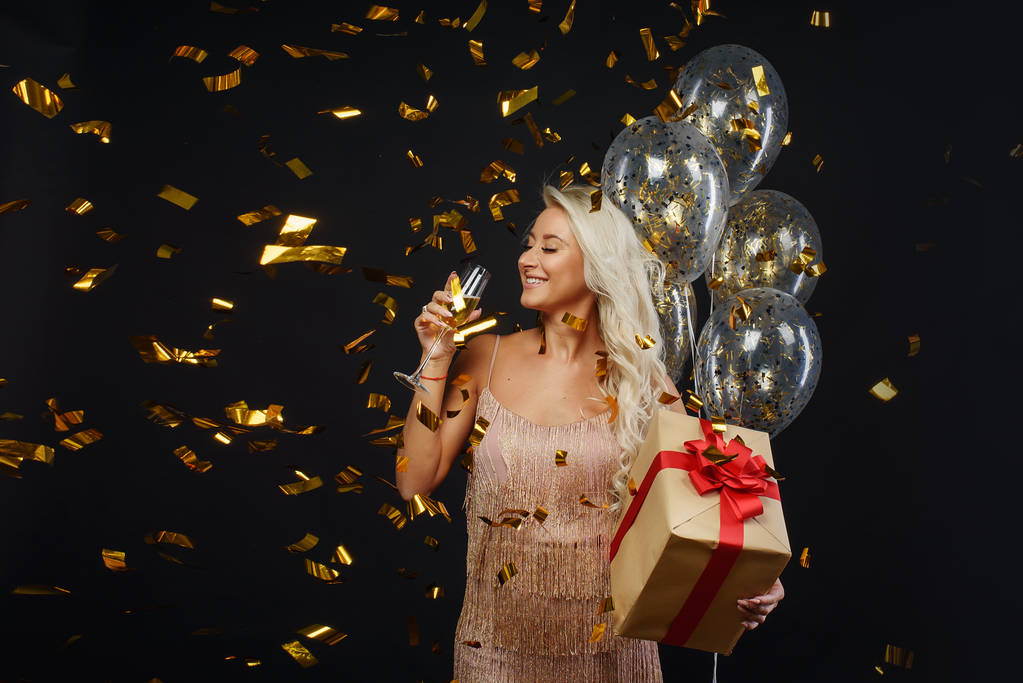 Luxus-örömteli fiatal nő ruhák ünneplése születésnap vagy karácsonyi buli konfetti a fekete háttér. Szórakozik, és mosolygott. A gazdaság jelenlegi, ajándék, golden lufi és a pohár pezsgő - Fotó, kép