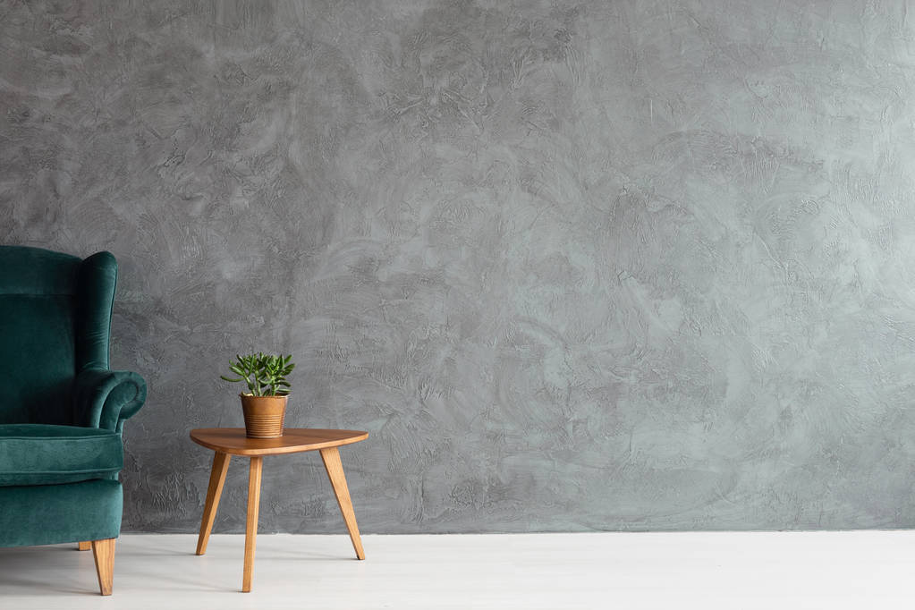 Donker groene velours fauteuil naast houten tafel met plant in pot, echte foto met kopie ruimte op lege betonnen wand - Foto, afbeelding