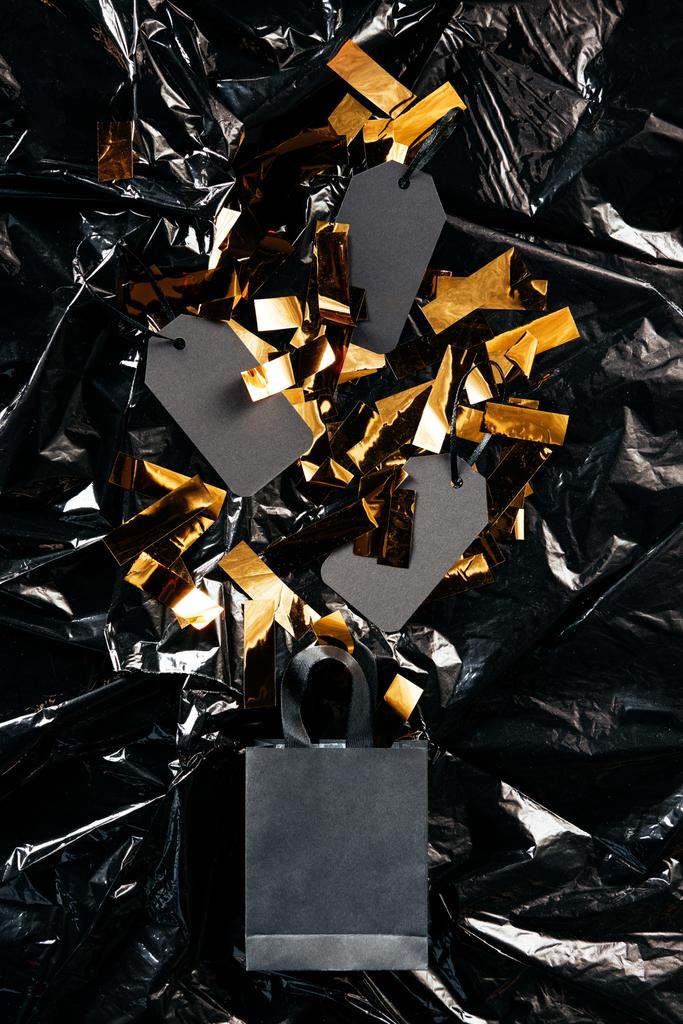 κάτοψη του χαρτί shopping τσάντα με κενή τιμή ετικέτες και χρυσές κομφετί αναδίπλωσης χαρτιού φόντο μαύρο, μαύρο Παρασκευή έννοια - Φωτογραφία, εικόνα