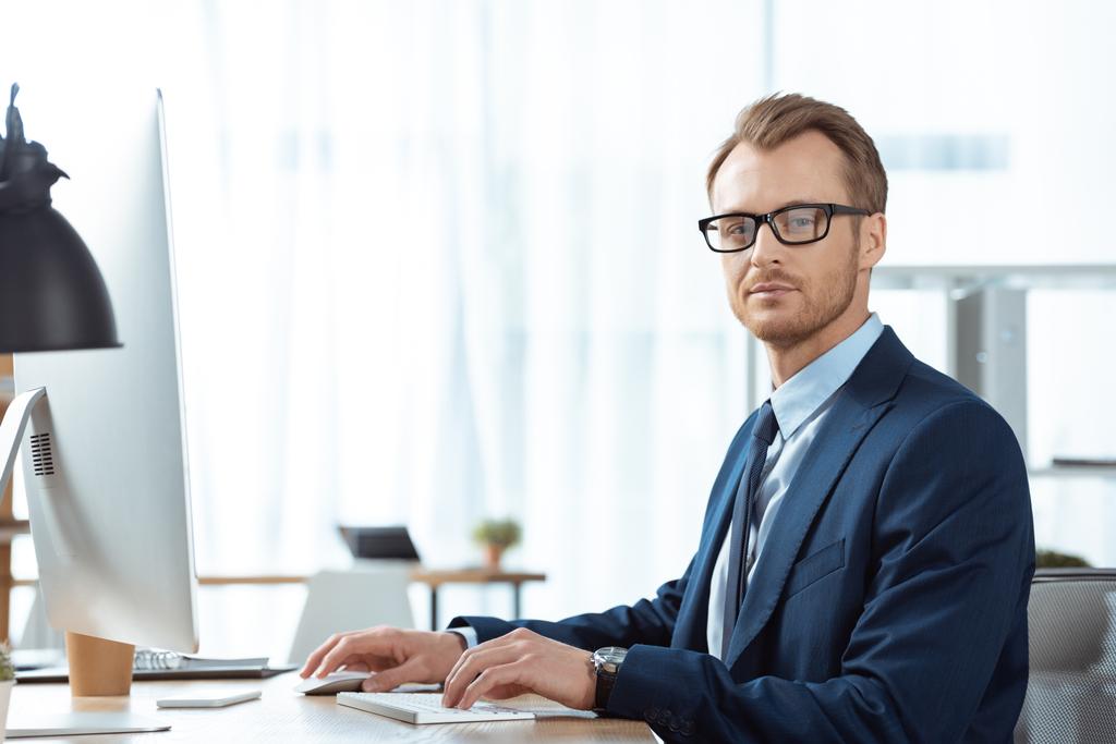 homem de negócios confiante em óculos olhando para a câmera e trabalhando à mesa com monitor de computador no escritório moderno
 - Foto, Imagem
