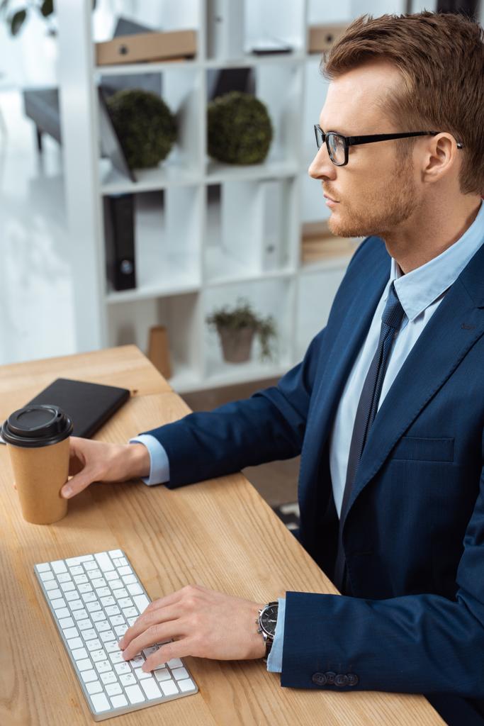 hombre de negocios serio en anteojos escribiendo en el teclado de la computadora en la mesa con la taza de café desechable en la oficina
  - Foto, Imagen