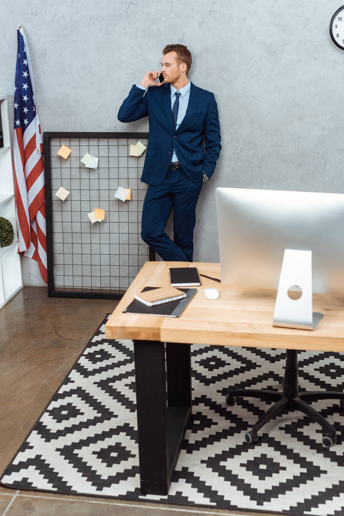 высокий угол обзора бизнесмена в костюме разговаривающего на смартфоне возле американского флага за стеной в современном офисе
  - Фото, изображение