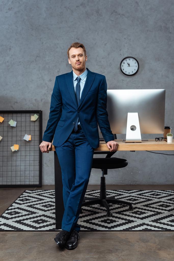 joyful young businessman in suit standing near table with computer monitor in office  - Photo, Image