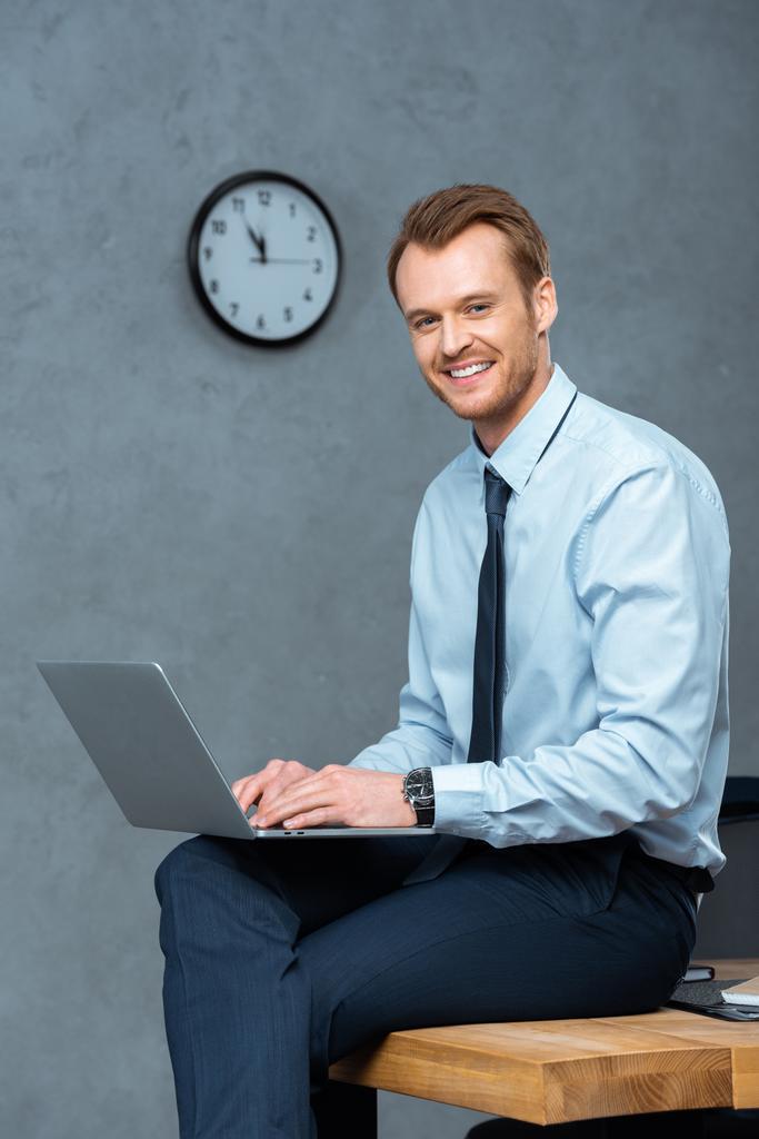jovem empresário feliz sentado na mesa e trabalhando no laptop no escritório moderno
  - Foto, Imagem