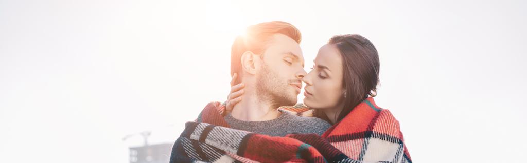 panoramic shot of beautiful young couple covering in plaid and kissing with sun shining behind - Photo, Image