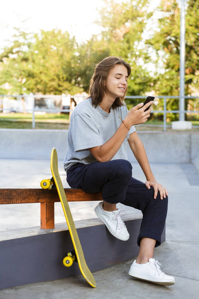 Image d'un jeune patineur joyeux assis dans le parc avec skateboard en utilisant un téléphone mobile
. - Photo, image
