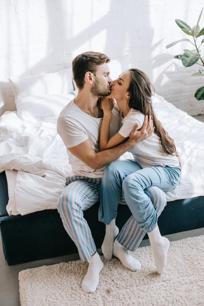 όμορφο νεαρό ζευγάρι στο πιτζάμες φιλιά ενώ κάθεται στο κρεβάτι πρωί - Φωτογραφία, εικόνα