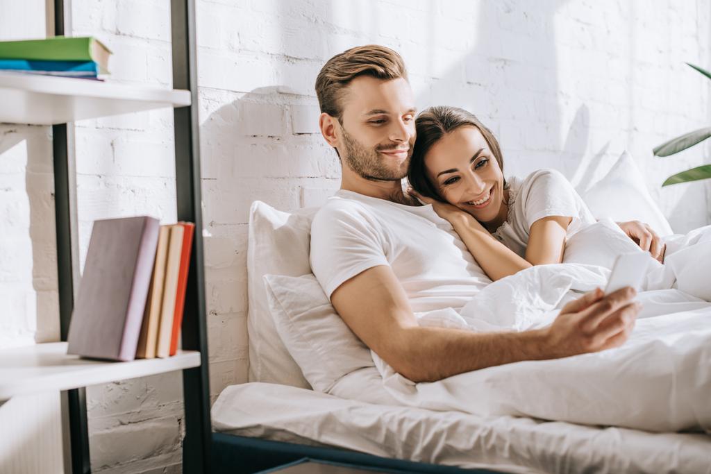 beautiful young couple relaxing in bed in morning and using smartphone - Photo, Image
