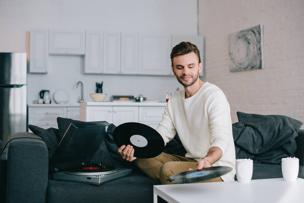 jovem atraente segurando discos para leitor de vinil enquanto sentado no sofá
 - Foto, Imagem