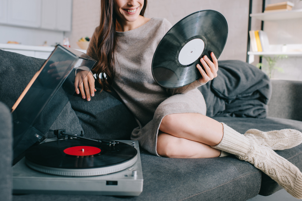 自宅のソファの上のビニール レコード プレーヤーで音楽を聴く若い女性のクロップ撮影 - 写真・画像