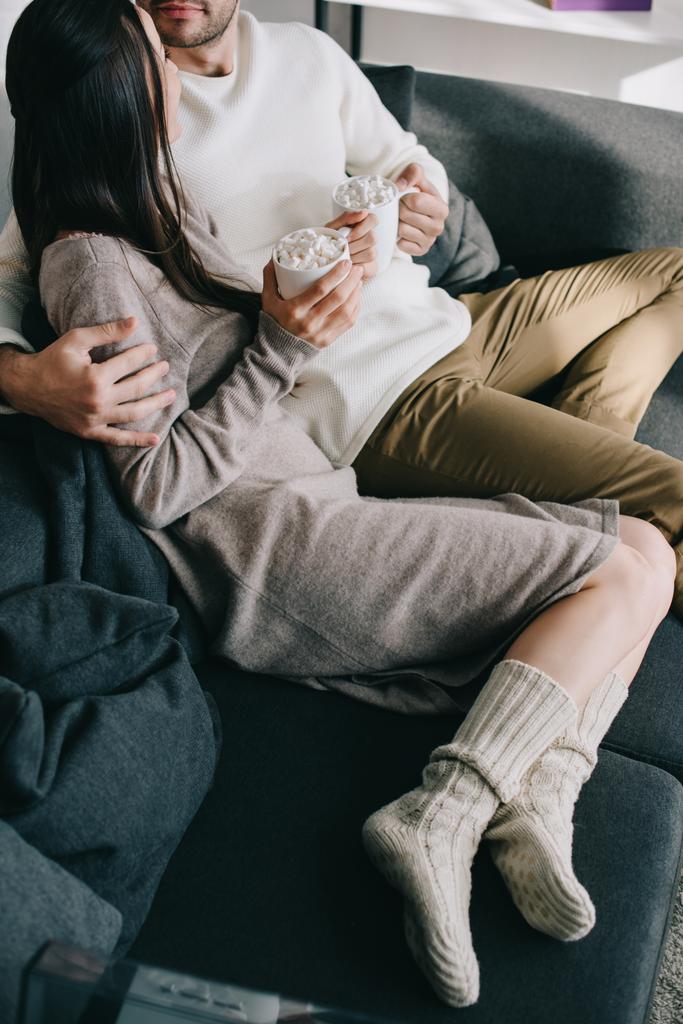 tiro recortado de pareja con tazas de cacao en calcetines de lana cálidos relajándose en el sofá en casa
 - Foto, imagen