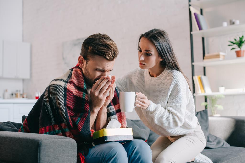 beautiful young woman holding cup of warming drink for sick boyfriend while he sneezing - Photo, Image