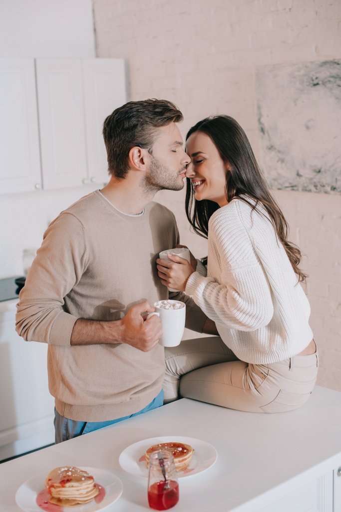beautiful young couple with cocoa and pancakes cuddling on kitchen - Photo, Image