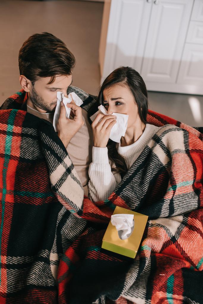 ill young couple with paper napkins sneezing while sitting on couch under plaid at home - Photo, Image