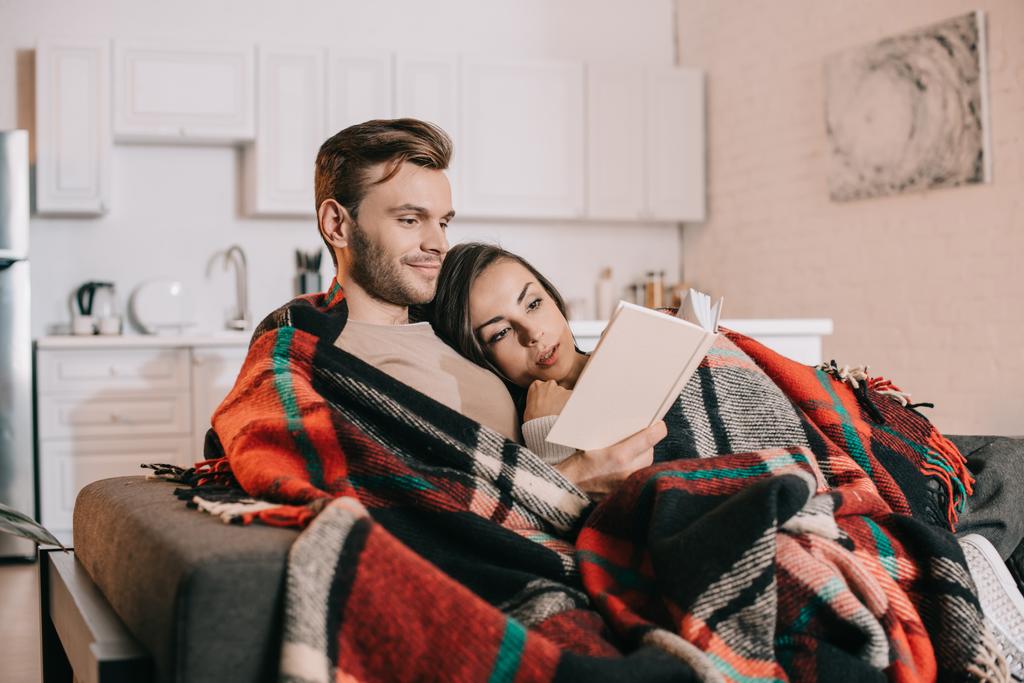 happy young couple reading book together on couch under plaid at home - Photo, Image