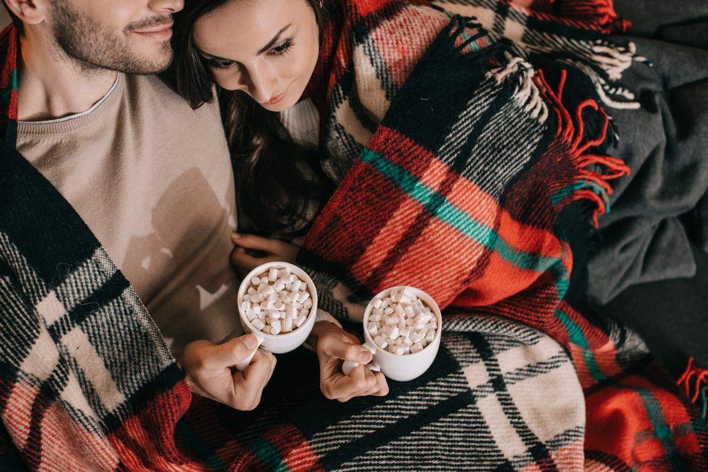 вид сверху на молодую пару с чашками какао с зефиром расслабляющий на диване под клеткой дома
 - Фото, изображение