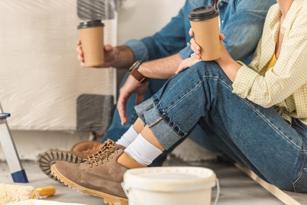 recortado disparo de joven pareja sosteniendo café para ir y sentarse en el suelo durante la mejora del hogar
 - Foto, Imagen