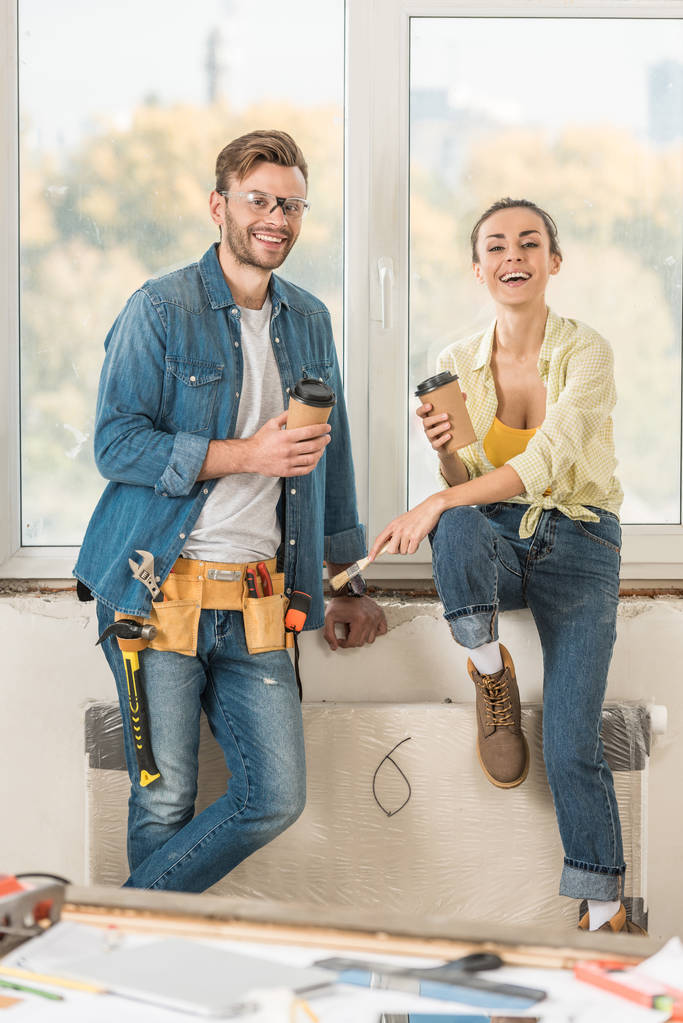 feliz joven pareja sosteniendo café para ir y sonriendo a la cámara durante la reparación de la casa
 - Foto, imagen