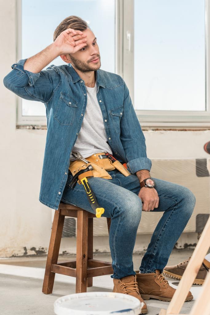 усталый молодой человек с поясом для инструментов сидит на стуле во время ремонта дома
 - Фото, изображение