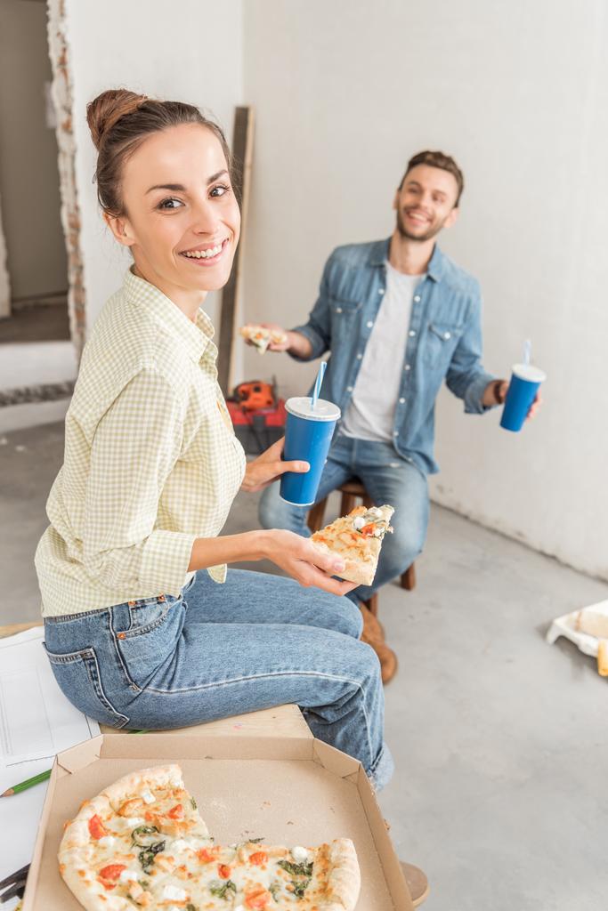 hoge hoekmening van gelukkig meisje glimlachend in de camera terwijl het eten van pizza met echtgenoot in nieuw appartement - Foto, afbeelding