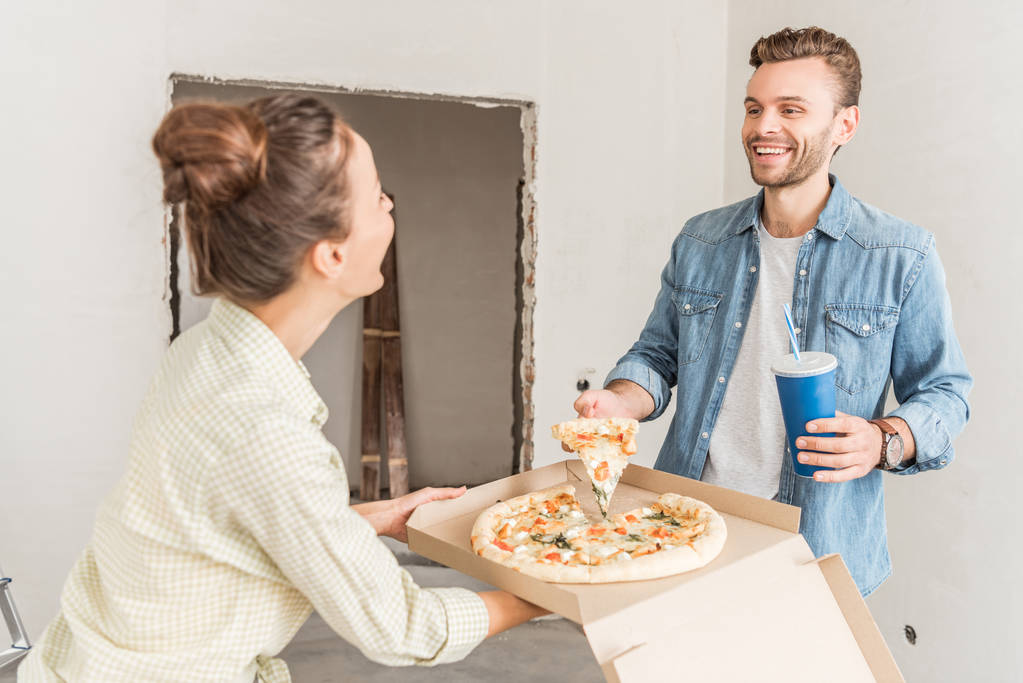 gelukkige jonge paar bedrijf doos met pizza en papier beker met verfrissende drank in nieuw appartement - Foto, afbeelding