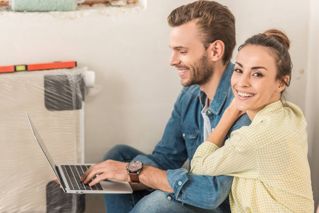 onnellinen nuori mies käyttää kannettavaa tietokonetta ja kaunis vaimo hymyilee kameralle uudessa talossa
 - Valokuva, kuva