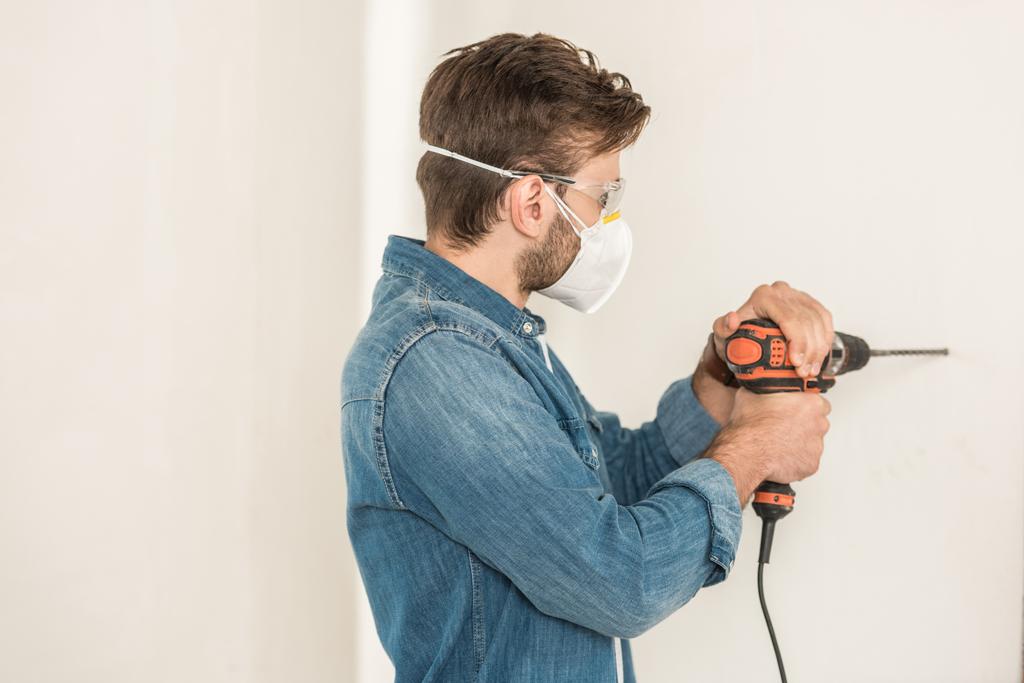 joven en ropa de trabajo protectora usando taladro eléctrico en la pared durante la reparación de la casa
  - Foto, imagen