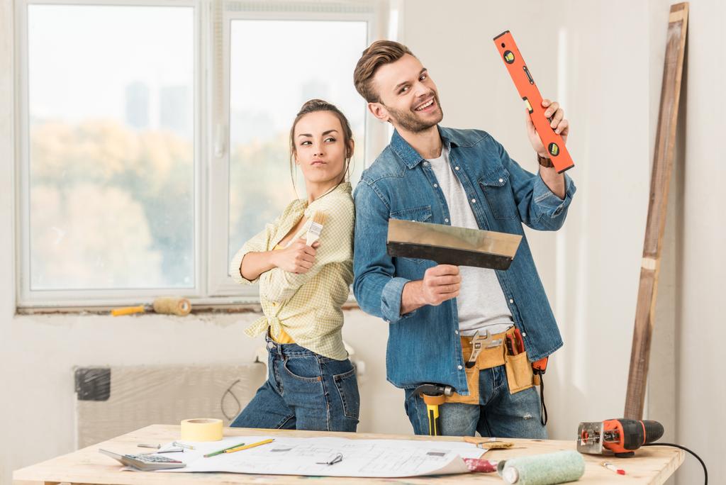 happy young couple holding renovation tools and smiling at camera in new apartment - Photo, Image