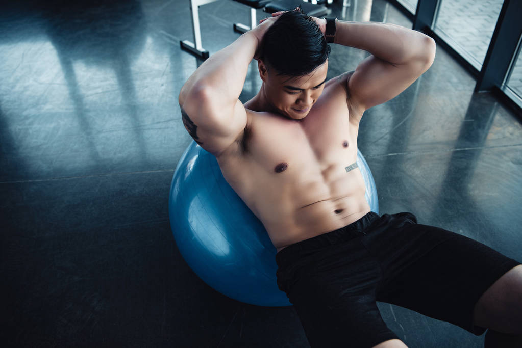 ジムでフィットネス ボールで腹筋のエクササイズをしている裸の胸に焦点を当てた若いアジア スポーツマン - 写真・画像