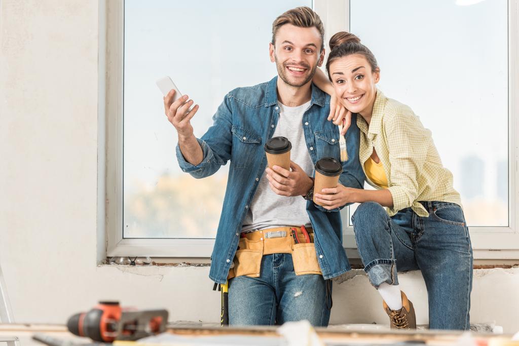happy young couple with coffee to go and smartphone smiling at camera during renovation - Photo, Image