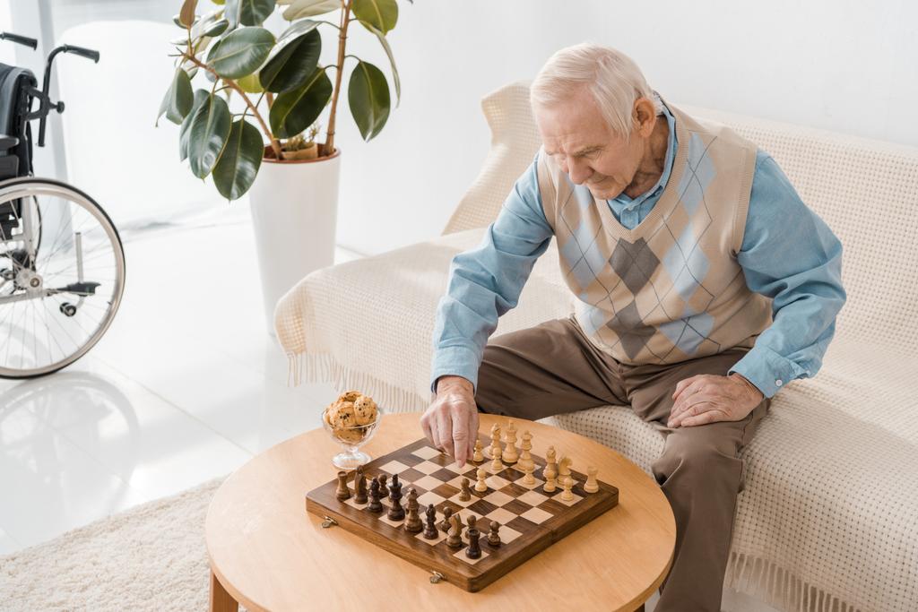 vanhempi mies istuu sohvalla ja pelaa shakkia
 - Valokuva, kuva
