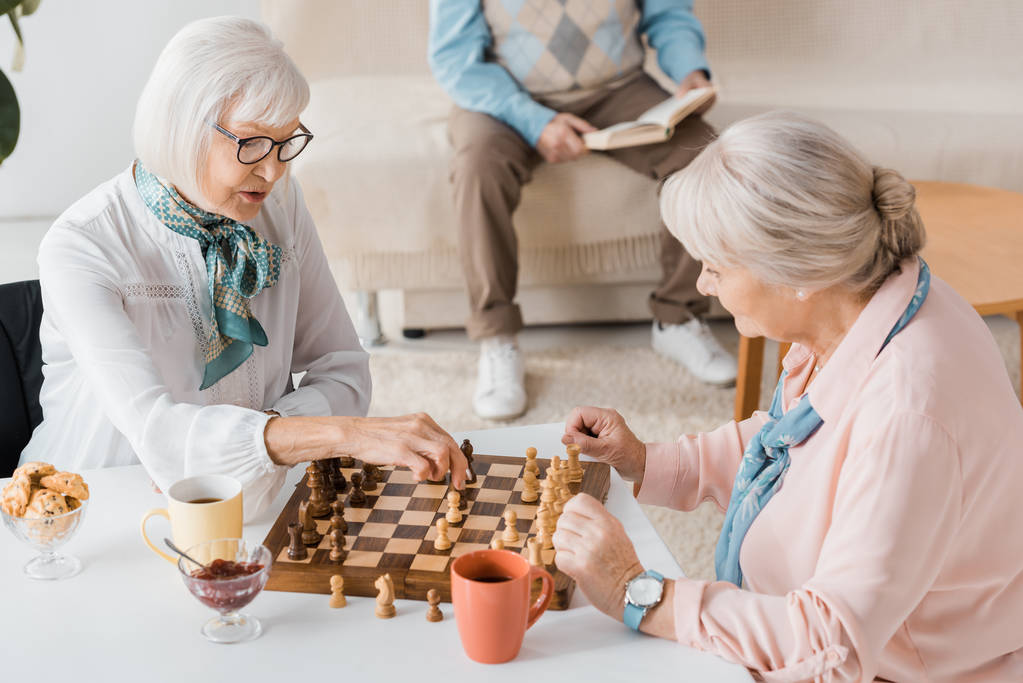 Senior vrouwen spelen schaak en drinken koffie terwijl man lezen boek op Bank - Foto, afbeelding