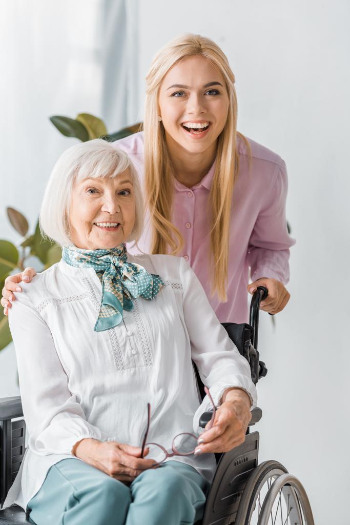 молодые и пожилые женщины в инвалидной коляске улыбаются и смотрят в камеру
 - Фото, изображение