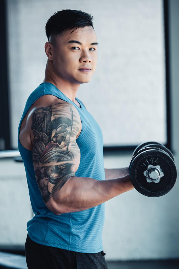 młody tatuażem asian sportowca, ćwiczenia z hantle w siłowni i patrząc na kamery - Zdjęcie, obraz